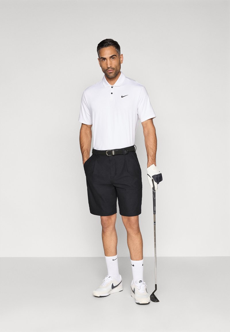 Polo blanc, short noir, ceinture noire, chaussettes blanches et gant de golf. Debout, tenant un club de golf. Chaussures de sport.