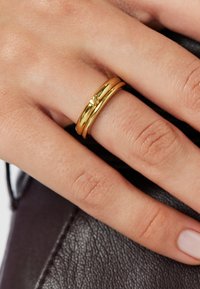 Dłoń z dwoma gładkimi złotymi pierścionkami na środkowym palcu, spoczywająca na ciemnej, teksturowanej skórzanej powierzchni.