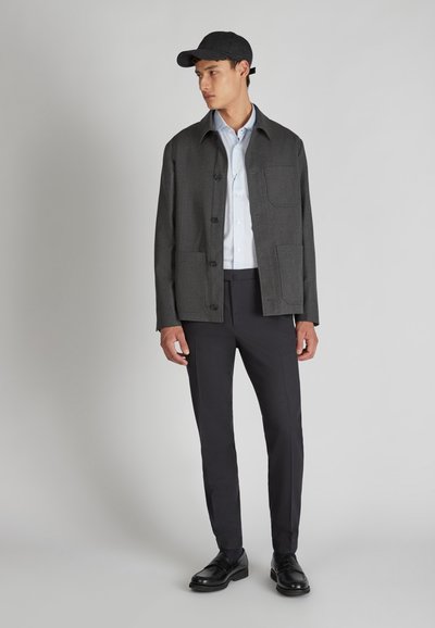 Giacca button-up in lana grigia con due tasche frontali, indossata sopra una camicia azzurro chiaro, abbinata a pantaloni scuri e scarpe nere. Berretto da baseball nero.