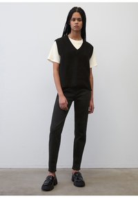 Svart strikket vest over en kremfarget t-skjorte, kombinert med mørkegrønne slim-fit bukser og kraftige svarte loafere. Enkelt design, teksturerte stoffer.