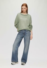 Hellgrüner, gestrickter Pullover mit lockerem Schnitt, kombiniert mit blauen, geraden Jeans. Model trägt silberne Sneakers.