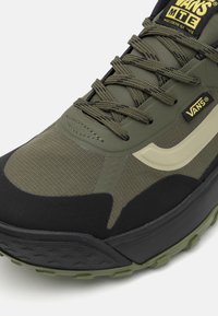 Chaussure de sport vert olive avec surface texturée, accents noirs et semelle extérieure en caoutchouc épaisse. Comprend un laçage renforcé et une étiquette de marque.