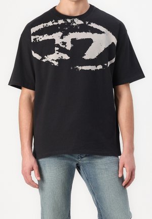 Homme portant un T-shirt noir oversize avec un graphisme abstrait blanc usé, associé à un jean bleu clair, debout devant un fond uni.