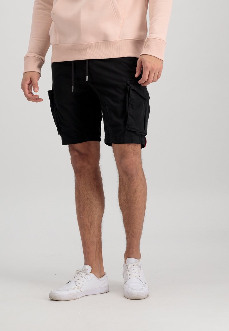 Shorts cargo noirs avec de multiples poches, taille élastique et fermeture à cordon. Portés avec des baskets blanches, sur un arrière-plan uni.