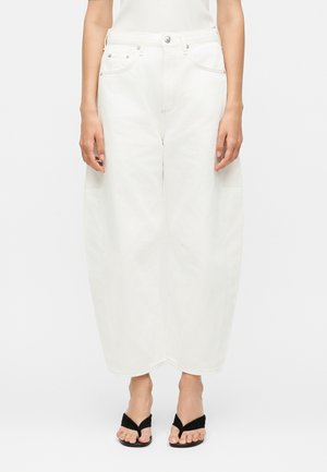 Kobieta ubrana w białe dżinsy z wysokim stanem i szerokimi nogawkami, połączone z czarnymi sandałami na obcasie z odkrytymi palcami, stojąca na jednolitym białym tle.