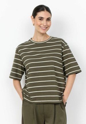 Femme aux cheveux foncés en queue de cheval souriante, portant un t-shirt à manches courtes rayé vert olive et blanc et un pantalon vert olive assorti, mains dans les poches.