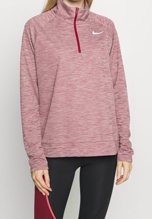 Femme portant un pull de sport à manches longues chiné rose avec une fermeture éclair au quart et des leggings noirs avec des panneaux bordeaux.
