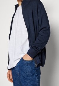 Man med marinblå dragkedja cardigan över en lös vit t-shirt med handen i fickan på blå denimjeans mot en enfärgad bakgrund.