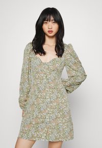 Vestido floral em verde, apresentando mangas bufantes, decote coração e fecho com botões. Os padrões incluem flores em azul claro e pêssego.
