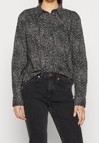 Musta napillinen paita, jossa on valkoisia epäsäännöllisiä pilkkuja, röyhelökaulus ja pitkät hihat, yhdistetty mustiin korkeavyötäröisiin farkkuihin.