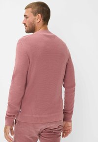 Mann, der nach links schaut, trägt einen strukturierten, langärmligen Pulli in rosarot sowie passende Cordhosen vor einem schlichten weißen Hintergrund.