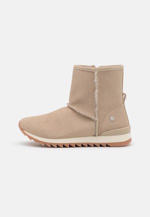 Beige ruskindet ankelstøvle med pelsforing, zigzag-gummisål og lille metallogo på baghæl.
