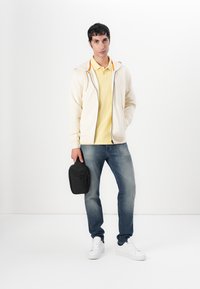 Lichtbeige zip-up hoodie met oranje accenten over een gele polo, gecombineerd met blauwe jeans en witte sneakers, terwijl hij een zwarte tas vasthoudt.