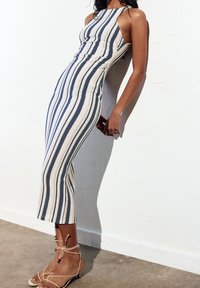 Robe midi rayée en beige et marine, avec une coupe ajustée, un dos nu et un tissu texturé. Associée à des sandales beiges à lacets.