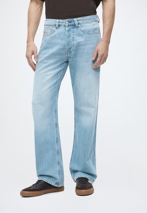 Homme portant un jean bleu clair ample et des baskets foncées avec des semelles marron, debout devant un fond blanc uni.