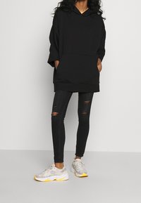 Sudadera negra de gran tamaño con capucha, que presenta un bolsillo frontal. Combinada con jeans skinny negros rasgados y zapatillas grises con acentos amarillos.