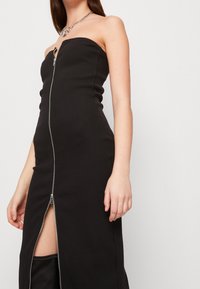 Schwarzes, geripptes, trägerloses Kleid mit einem Frontreißverschluss und einem Schlitz, mit glatter Textur und figurbetontem Design. Akzentuiert mit einer Kettenhalskette.