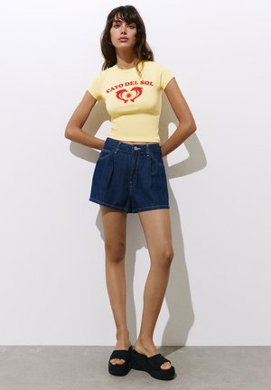 Femme debout, les mains derrière le dos, portant un t-shirt jaune "Cayo Del Sol", un short en jean bleu foncé et des sandales compensées noires.