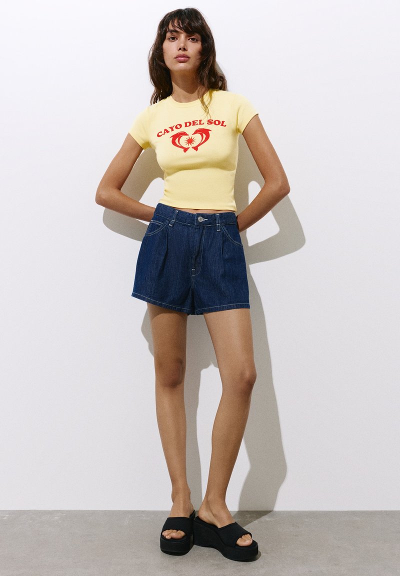 Femme debout, les mains derrière le dos, portant un t-shirt jaune "Cayo Del Sol", un short en jean bleu foncé et des sandales compensées noires.