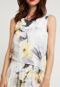 Kwiatowa bezrękawnik z warstwowym wzorem. Posiada jasnoszare i żółte kwiaty na białym tle, delikatne plisowania i lekką tkaninę.