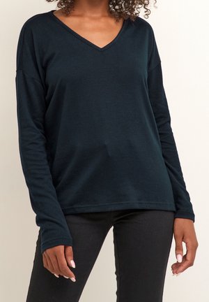 Maglietta verde scuro a maniche lunghe con scollatura a V, realizzata in tessuto morbido. Presenta una vestibilità rilassata e spalle cadenti. Abbinata a pantaloni neri, mani visibili.