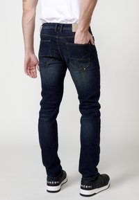 Jeans in denim scuro con vestibilità slim. Presentano due tasche posteriori, cuciture a contrasto e un'etichetta del logo sulla vita. Indossati con scarpe da ginnastica nere.