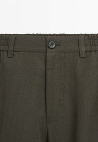 Mörkgröna byxor med elastisk midjeresår, bältesöglor och en svart knapp som stängning framtill i mitten.