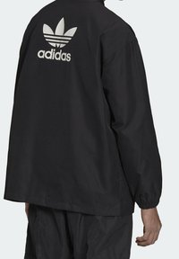 Svart vadderad jacka med långa ärmar, som har en stor vit Adidas trefoil-logga på baksidan och elastiska ärmslut för en tät passform.