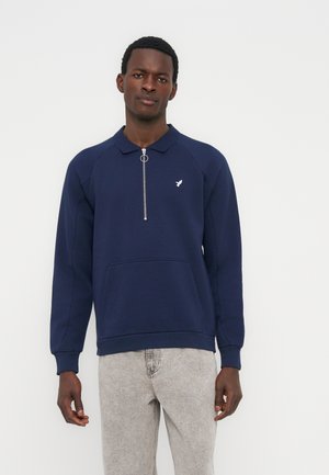 Homme portant un sweat-shirt zippé bleu marine avec un petit logo blanc sur la poitrine et un jean gris clair, debout devant un fond blanc.