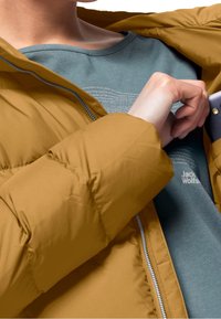 Senapsgul puffjacka med dragkedja, med en mjuk, vadderad textur. Inuti visar en blå-grön skjorta subtil text och en logotyp.