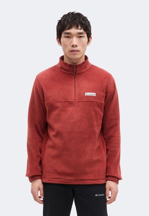 Homme portant un pull polaire Columbia couleur rouille avec une fermeture éclair quart de zip et un pantalon noir Columbia, faisant face, devant un fond uni.