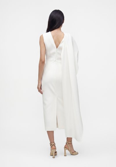 Femme aux longs cheveux foncés portant une robe midi blanche sans manches avec un drapé asymétrique à l'épaule et des sandales à talons beiges, de dos.