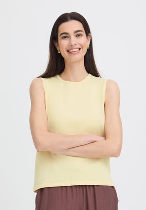 Usmievavá žena s dlhými tmavými vlasmi má na sebe žltý bezrukávový top a zlaté náušnice, stojí so založenými rukami na bielom pozadí.