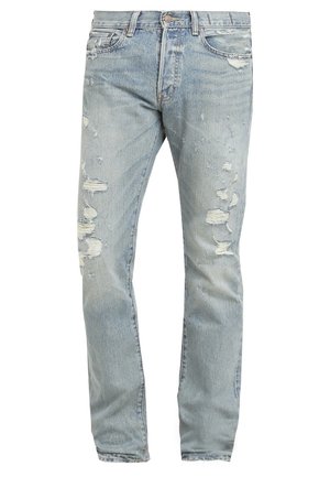 Uomo con la barba che indossa una giacca di pelle marrone, una t-shirt grigio chiaro, jeans azzurri strappati e sneakers nere, in piedi con una gamba incrociata.