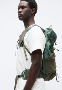 Σακίδιο πλάτης σε σκούρο πράσινο και ελιά ύφασμα με υφασμάτινα πάνελ, ρυθμιζόμενους ιμάντες και πλαϊνές τσέπες. Διαθέτει ρυθμιζόμενους ιμάντες στο στήθος και τη μέση.