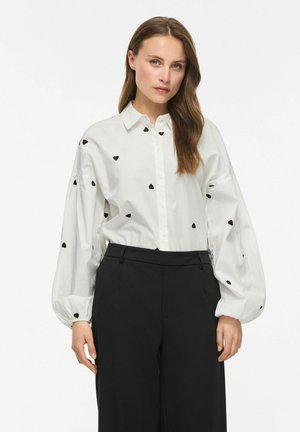 Femme portant une chemise blanche à boutons avec des motifs de cœurs noirs et un pantalon noir à jambes larges, debout devant un fond blanc uni.