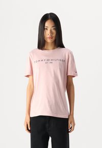 Lys rosa bomulls T-skjorte med korte ermer og rund hals, med "TOMMY HILFIGER"-logoen trykket i marineblått på framsiden.