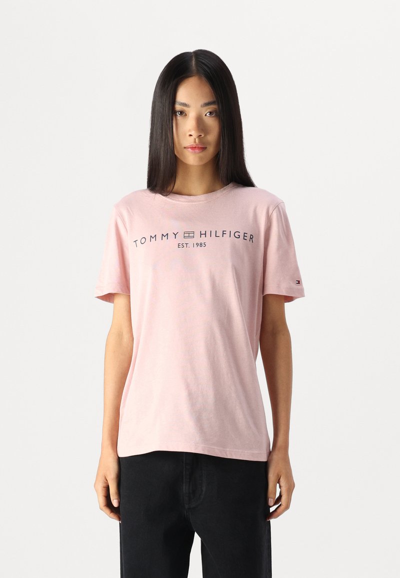Lys rosa bomulls T-skjorte med korte ermer og rund hals, med "TOMMY HILFIGER"-logoen trykket i marineblått på framsiden.