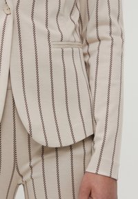 Beżowa marynarka w pionowe brązowe paski, wykonana z gładkiej tkaniny. Posiada pojedyncze zapięcie na guzik oraz boczną kieszeń.