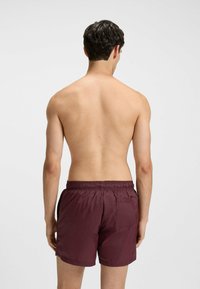 Bordowe kąpielówki z elastycznym pasem, bocznymi kieszeniami i jedną tylną kieszenią. Lekki, gładki materiał. Pokazano widok z tyłu.