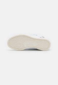 Bílé tenisky s texturovanou gumovou podešví, která má hexagonální vzor trakce a malé logo Reebok na boku. Minimalistický design.