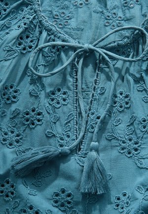 Hele sinine tikitud pluus, millel on lillemustrid, seotav detail ja pitsid. Pehme kangas tekstuursete õmbluste ja välja lõigatud aktsentidega.