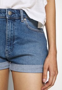 Jeansshorts i medelblått med hög midja, upprullade fållar och metallknappstängning. Har kontrasterande sömmar och fem fickor.