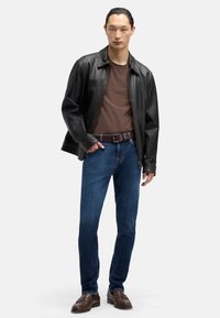 Czarna skórzana kurtka, brązowy t-shirt, niebieskie dżinsy, brązowy pasek i ciemnobrązowe mokasyny. Model stoi z jedną ręką w kieszeni.