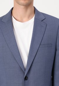 Jasnoniebieska marynarka wykonana z teksturowanej tkaniny, z klapami, z pojedynczym zapięciem na guzik i kieszenią, noszona na białym T-shircie.