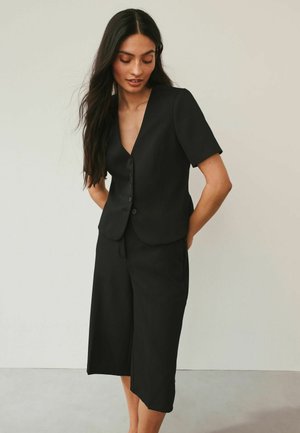 Woman with long dark hair wearing a black short-sleeve buttoned blazer and asymmetrical black midi skirt, standing against a plain background.