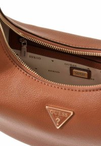 Interieur van een bruine leren handtas met ritssluiting, voorzien van een "GUESS" logo label en een lichtgekleurde voering met een herhaald merkpatroon.