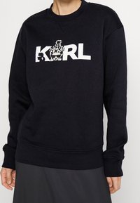 Felpa nera con maniche lunghe, polsini e orlo a coste. Presenta una grafica bianca "KARL" con un design giocoso sopra il testo.