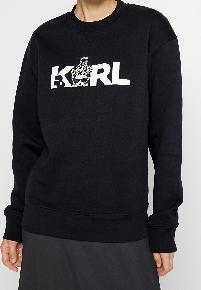 Felpa nera con maniche lunghe, polsini e orlo a coste. Presenta una grafica bianca "KARL" con un design giocoso sopra il testo.