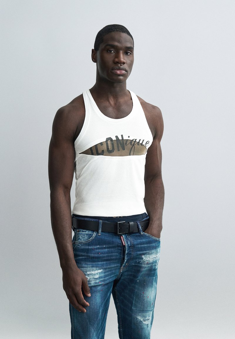 Uomo muscoloso che indossa una canottiera bianca con ritaglio e scritta "ICONique", abbinata a jeans blu strappati e cintura nera, in piedi su sfondo grigio.